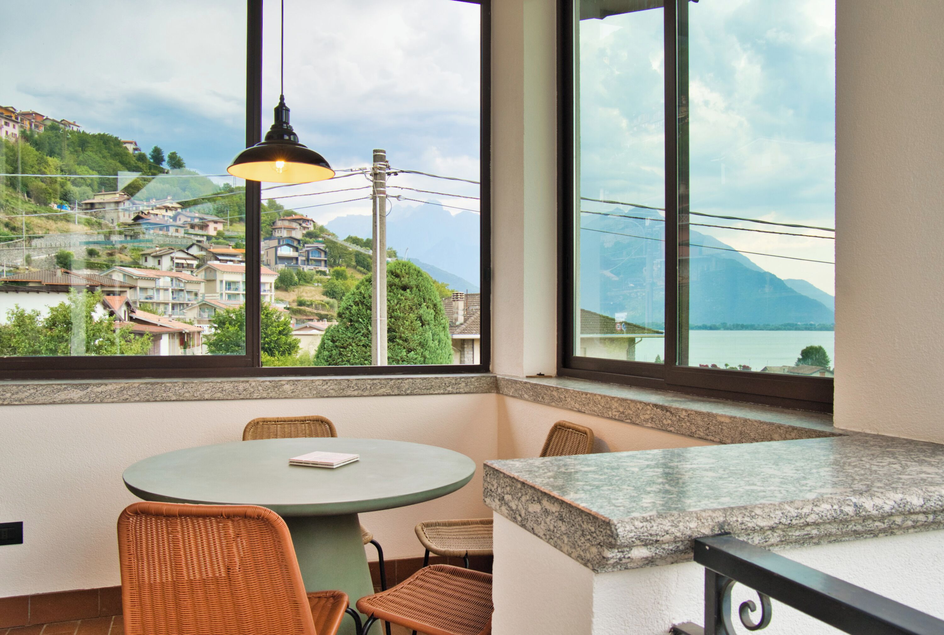 A corner of a room with a round table, wicker chairs, and a view of a hillside village through large windows.