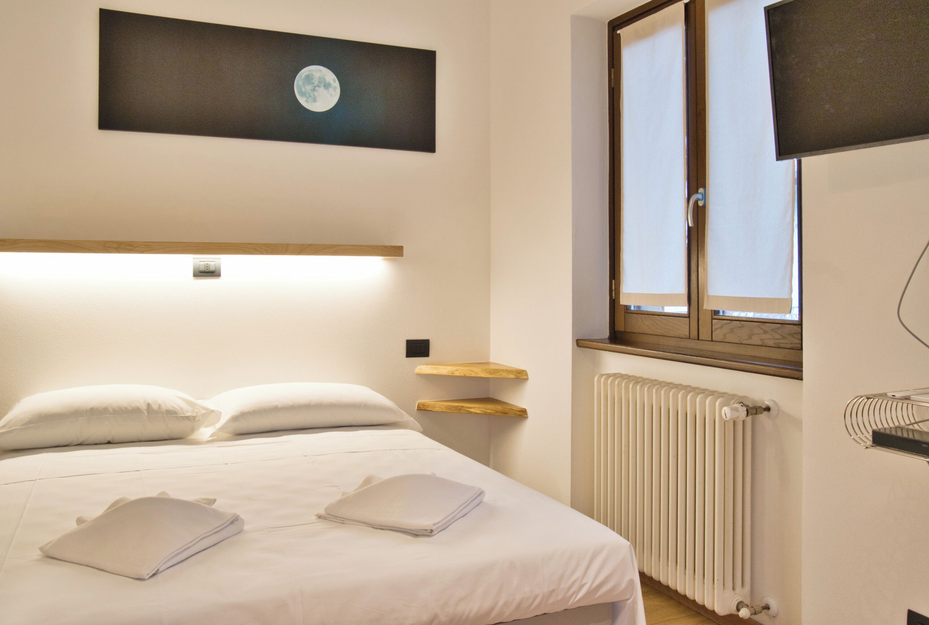 Een net opgemaakt bed met kussens en opgevouwen handdoeken, een wandplank, een raam met uitzicht, en een radiator.