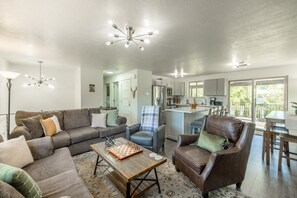 Ein modernes Wohnzimmer mit einer Couch, einem Sessel und einem Couchtisch, eine Küche mit Kochinsel sowie ein Balkon mit Tisch.