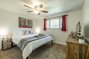 Ein Schlafzimmer mit einem Bett, einem Deckenventilator, einem Fenster mit Vorhängen, einem Nachttisch mit Lampe und einer Kommode mit Pflanze.