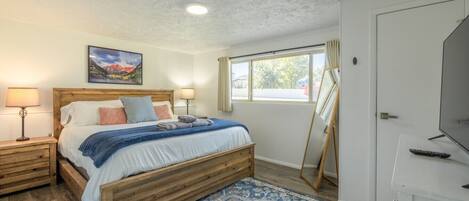Un dormitorio con una cama de madera, lámparas de noche, un cuadro, un ventanal con cortinas, un televisor y una alfombra estampada.