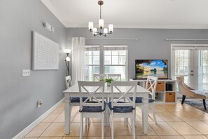 Un espace de salle à manger moderne avec une table et des chaises blanches, un lustre et une grande fenêtre avec des rideaux.
