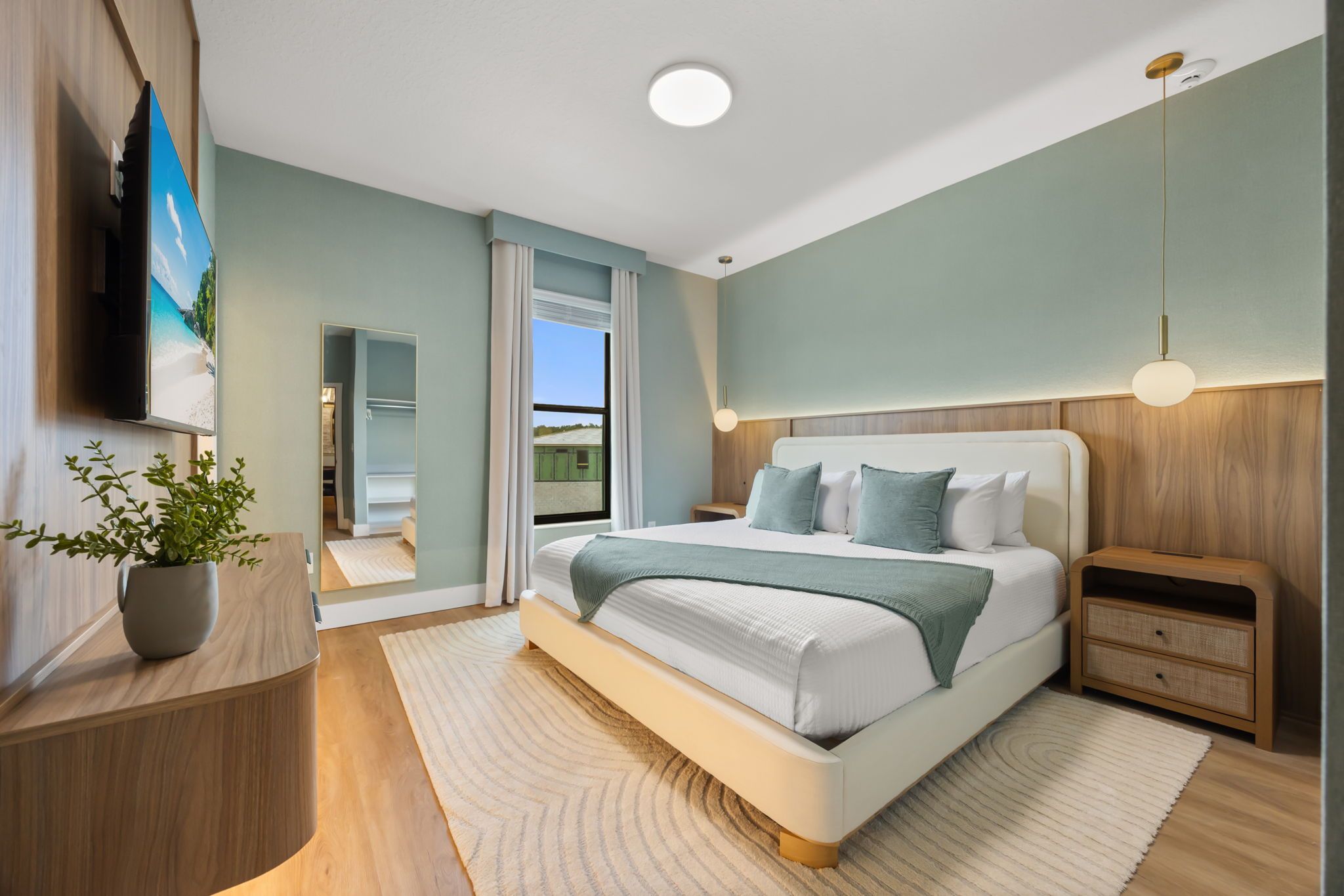 A modern bedroom with a large bed, bedside tables, a TV, and a view of a green landscape.