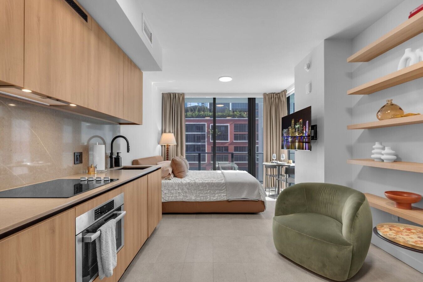 A modern hotel room with a kitchenette, a bed with a city view, a green armchair, and a TV on the wall.