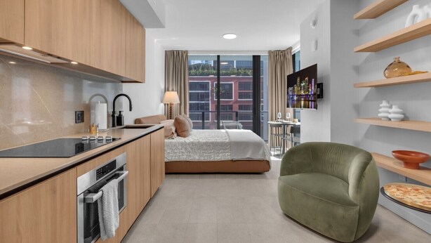 Quarto de hotel moderno com cozinha compacta, cama com vista para a cidade, poltrona verde e televisão na parede.