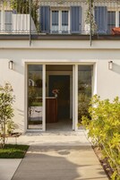 Ein modernes Gebäude mit Glastür, einer kleinen Außenbestuhlung und einem Balkon mit blauen Rollläden.