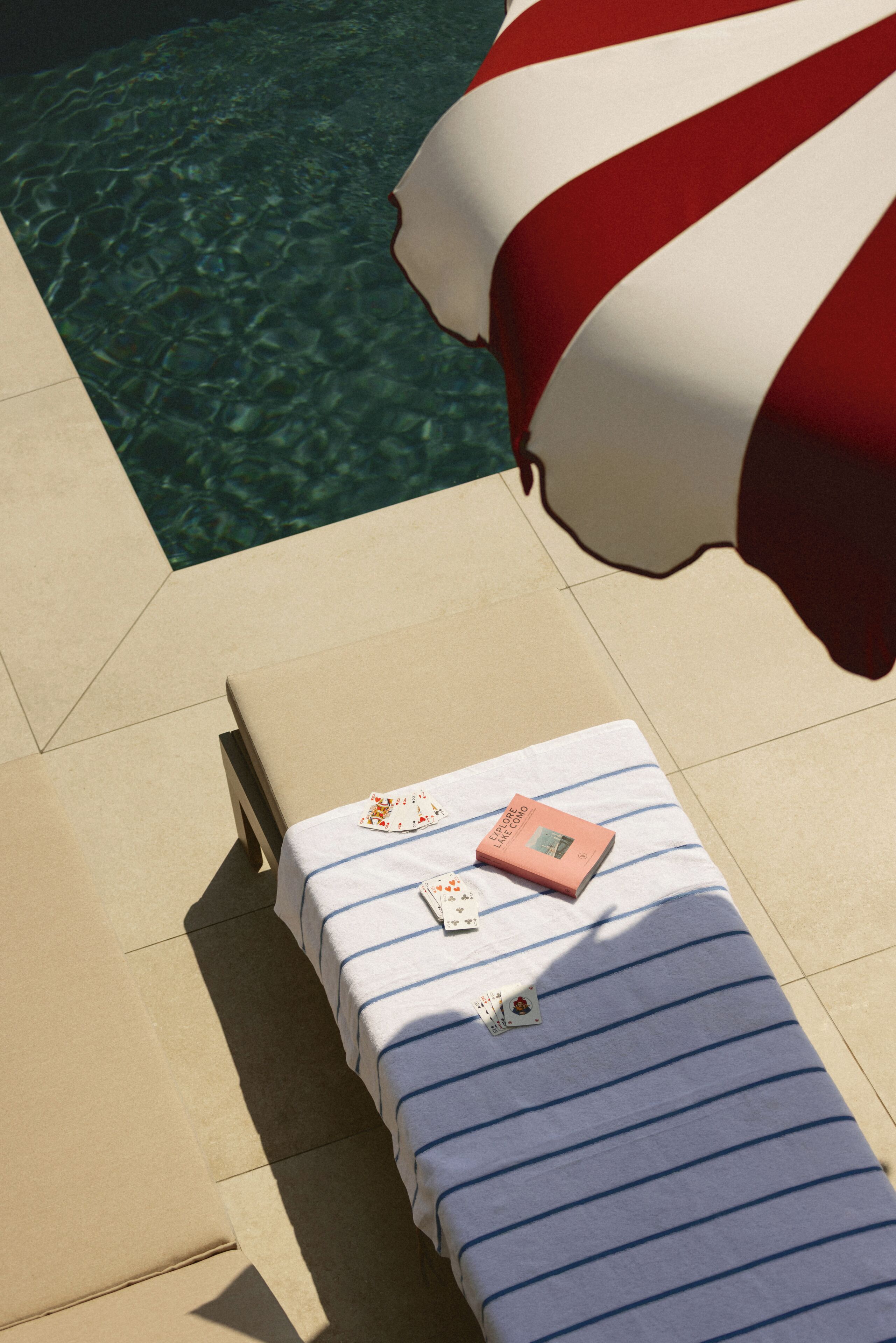 Un lettino con biancheria a righe, un libro intitolato 'Esplora il Lago di Como' e un mazzo di carte su un pavimento piastrellato vicino a una piscina.
