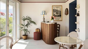 Una habitación con un escritorio de madera, una mesa de vidrio, una planta en maceta, un otoman a rayas y un cuadro en la pared.