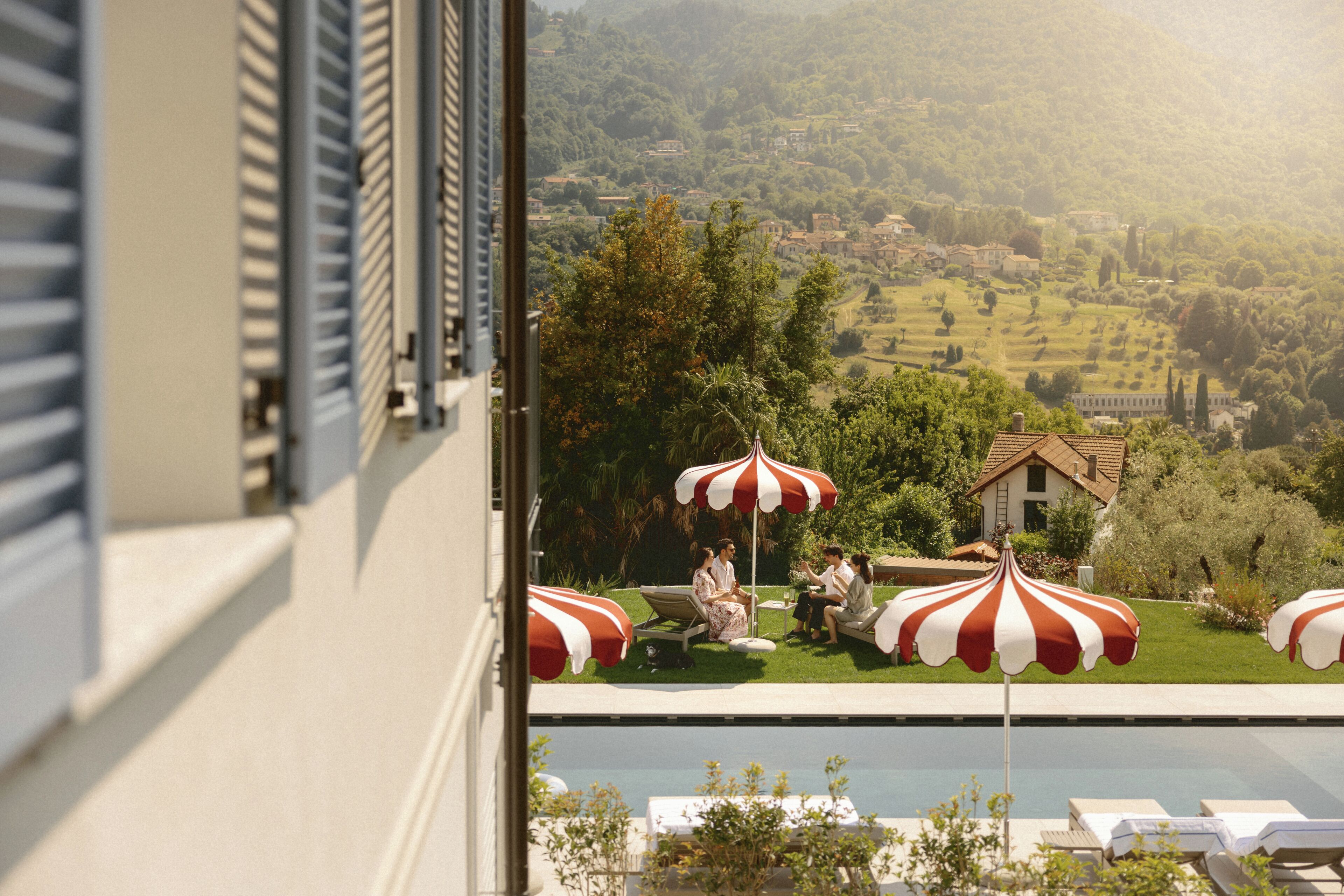 A poolside area with lounge chairs and umbrellas, a view of a hillside village, and a building with shutters.