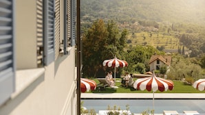 Zona junto a la piscina con sillones y sombrillas, vista a un pueblo en la ladera, y un edificio con persianas.