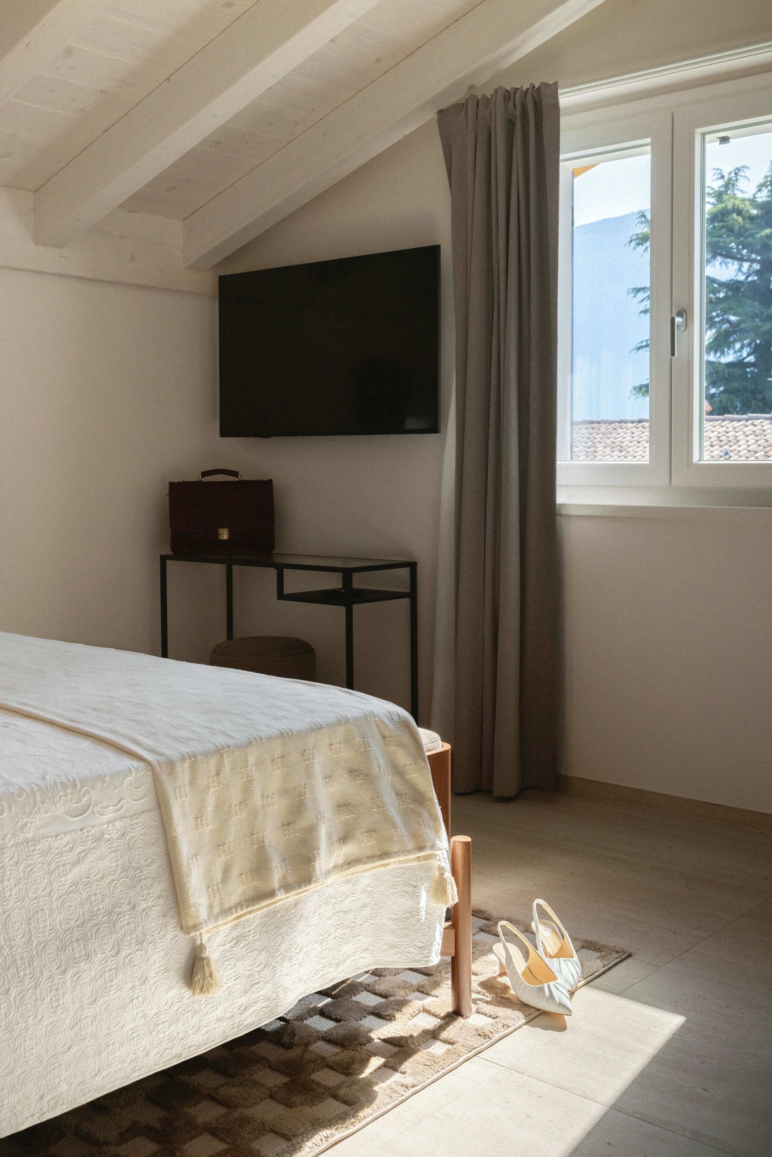 A bedroom with a bed, a TV mounted on the wall, a window with curtains, and a small table with a chair.