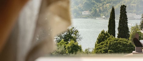 A hand adjusting a window blind, revealing a view of a lake, trees, and a distant building.