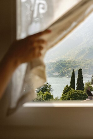 A hand adjusting a window blind, revealing a view of a lake, trees, and a distant building. - Hortus Bellagio (Bellagio)