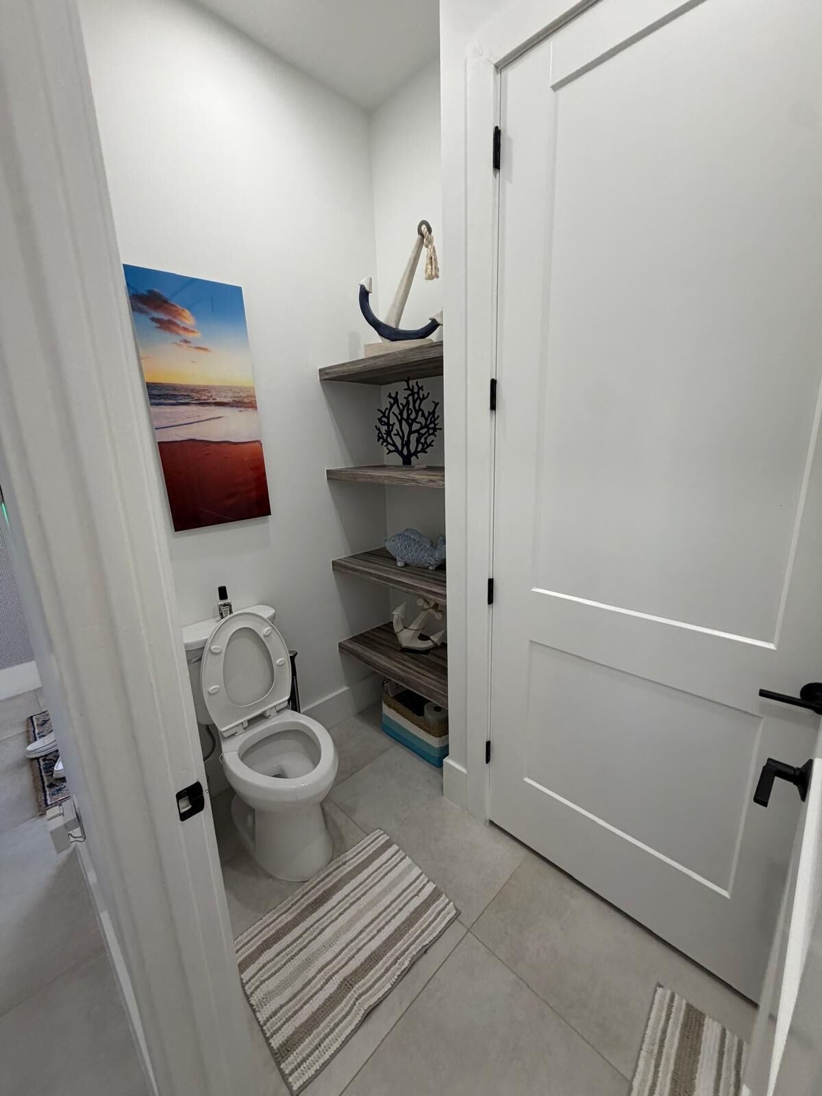 A bathroom with a toilet, a shelf with decorative items, and a painting on the wall.