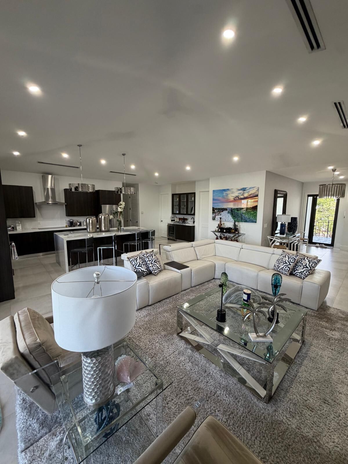 A modern living room with a glass coffee table, a sofa set, and a dining area with a kitchen in the background.