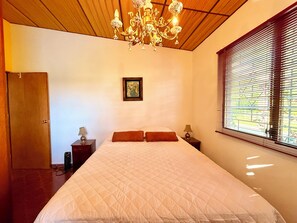 Ein Schlafzimmer mit einem großen Bett, einer Holzbalkendecke, einem Kronleuchter, einem Fenster mit Jalousien und einer Tür.