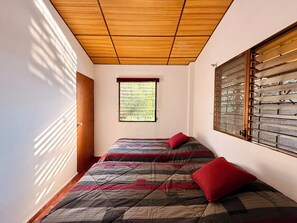 A room with a sloped ceiling, a window with blinds, a bed with a patterned bedspread, and a door. - Mountain Retreat among Coffee fields & Nature (Los Naranjos)