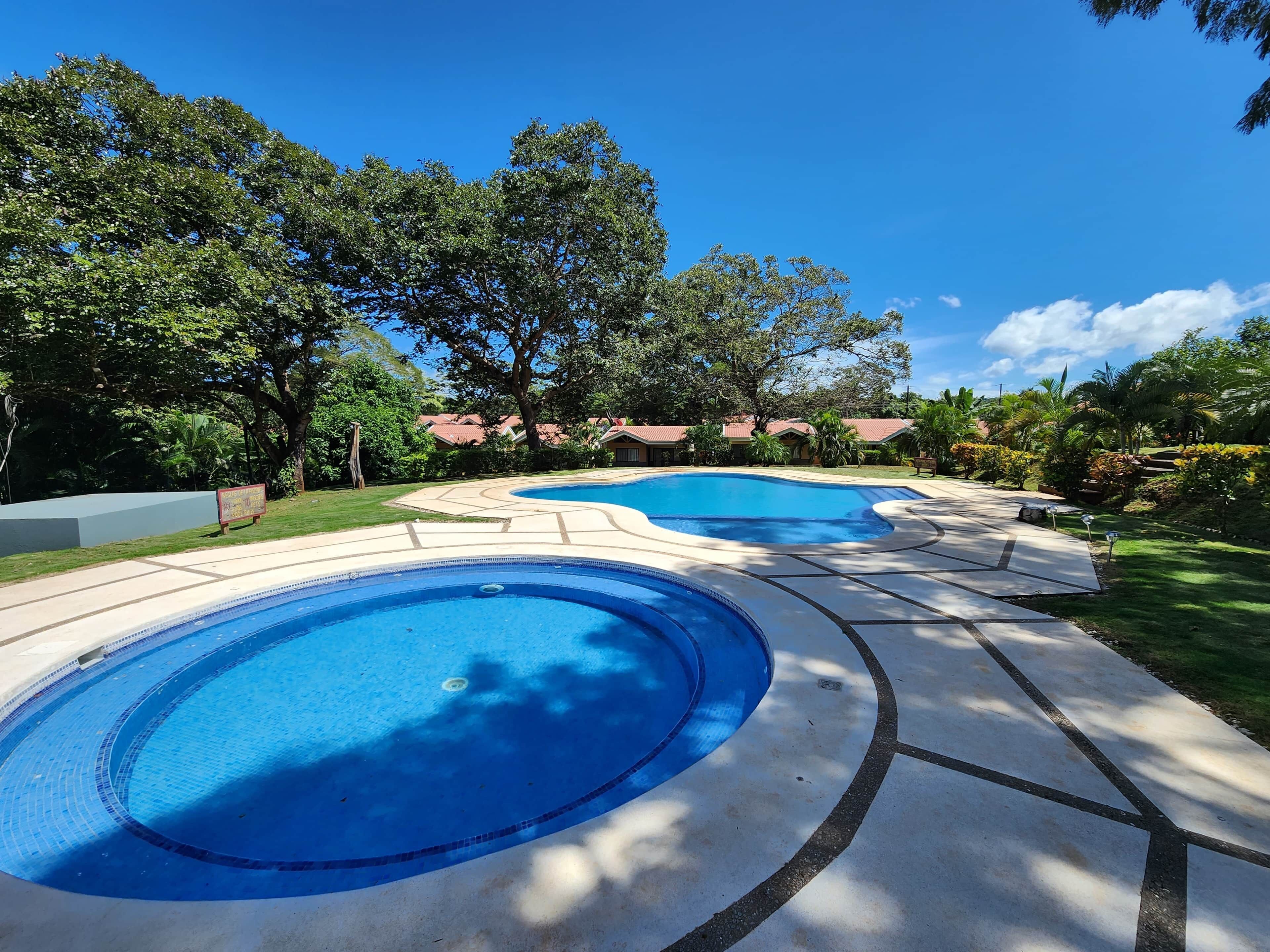 Une piscine dont le bord est recouvert de carrelage, entourée d’une terrasse en béton, d’arbres et sous un ciel bleu dégagé.