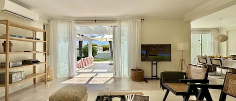 A spacious living area with a sofa, coffee table, and a dining table with chairs. There is a bookshelf, a television, and a view of the outdoors through a glass door.