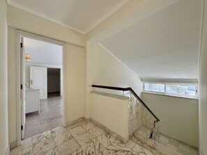 Un escalier avec un sol en marbre menant à un niveau supérieur, une porte donnant sur une pièce avec un lit, et une fenêtre ornée de balustrades en fer forgé.
