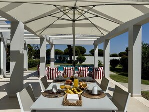 Un espace extérieur abrité, aménagé pour les repas, avec des tables préparées pour le petit-déjeuner, et offrant une vue sur une piscine.