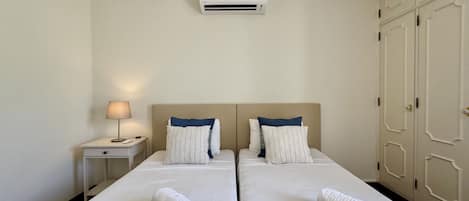 A hotel room with two beds, a nightstand, a lamp, an air conditioning unit, and a wardrobe.