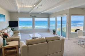 A spacious living room with a large window overlooking the ocean, a beige sofa, a coffee table, and a flat-screen TV mounted on the wall.