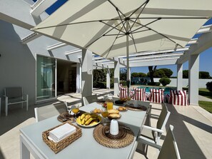 Un patio al aire libre con una mesa lista para el desayuno, una piscina al fondo y una zona de estar cubierta.