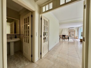 A hallway with a bathroom on the left, a dining area with chairs and a table in the center, and a fireplace with a bookshelf in the background. - VILAMOURA SUN & BEACH VILLA WITH POOL by HOMING (Vilamoura)