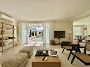 A spacious living area with a sofa, coffee table, and a dining table with chairs. There is a bookshelf, a television, and a view of the outdoors through a glass door. - VILAMOURA SUN & BEACH VILLA WITH POOL by HOMING (Vilamoura)