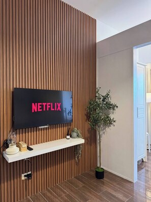 Ein modernes Wohnzimmer mit einer Holzpaneelwand, einem montierten Fernseher, der 'NETFLIX' anzeigt, einem weißen Regal mit Dekorationen und einer Topfpflanze.