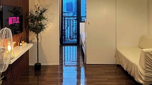 Quarto de hotel com piso de madeira, uma cama, mesa de cabeceira com ventilador, planta em vaso e televisão na parede.