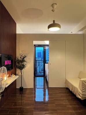 Ein Hotelzimmer mit Holzboden, einem Bett, einem Nachttisch mit Ventilator, einer Topfpflanze und einem an der Wand befestigten Fernseher.