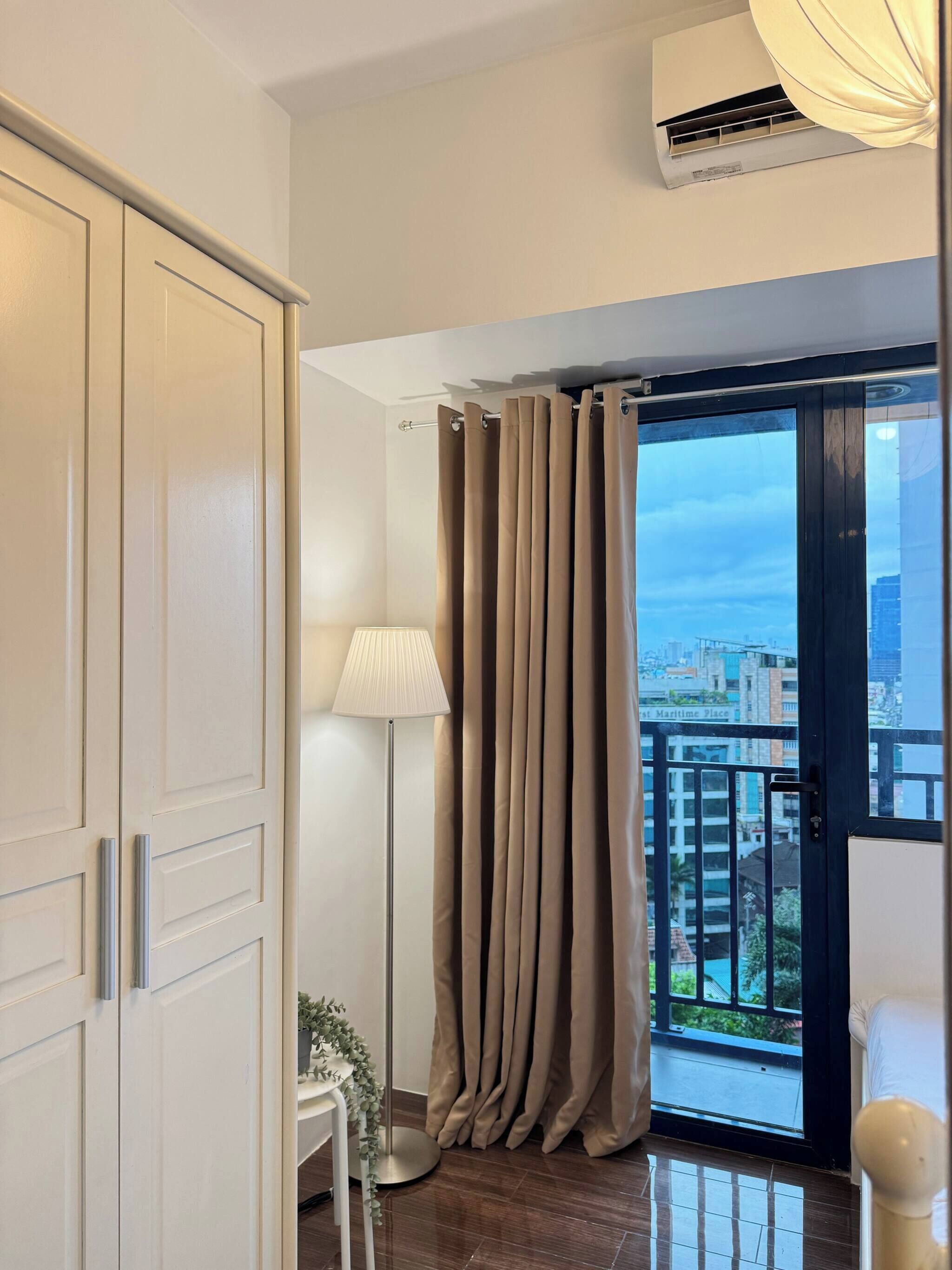 A room with a sliding door to a balcony, a standing lamp, and a view of a cityscape.