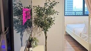 Quarto de hotel moderno com uma cama, televisão, planta em vaso e vista para a cidade através de uma janela.