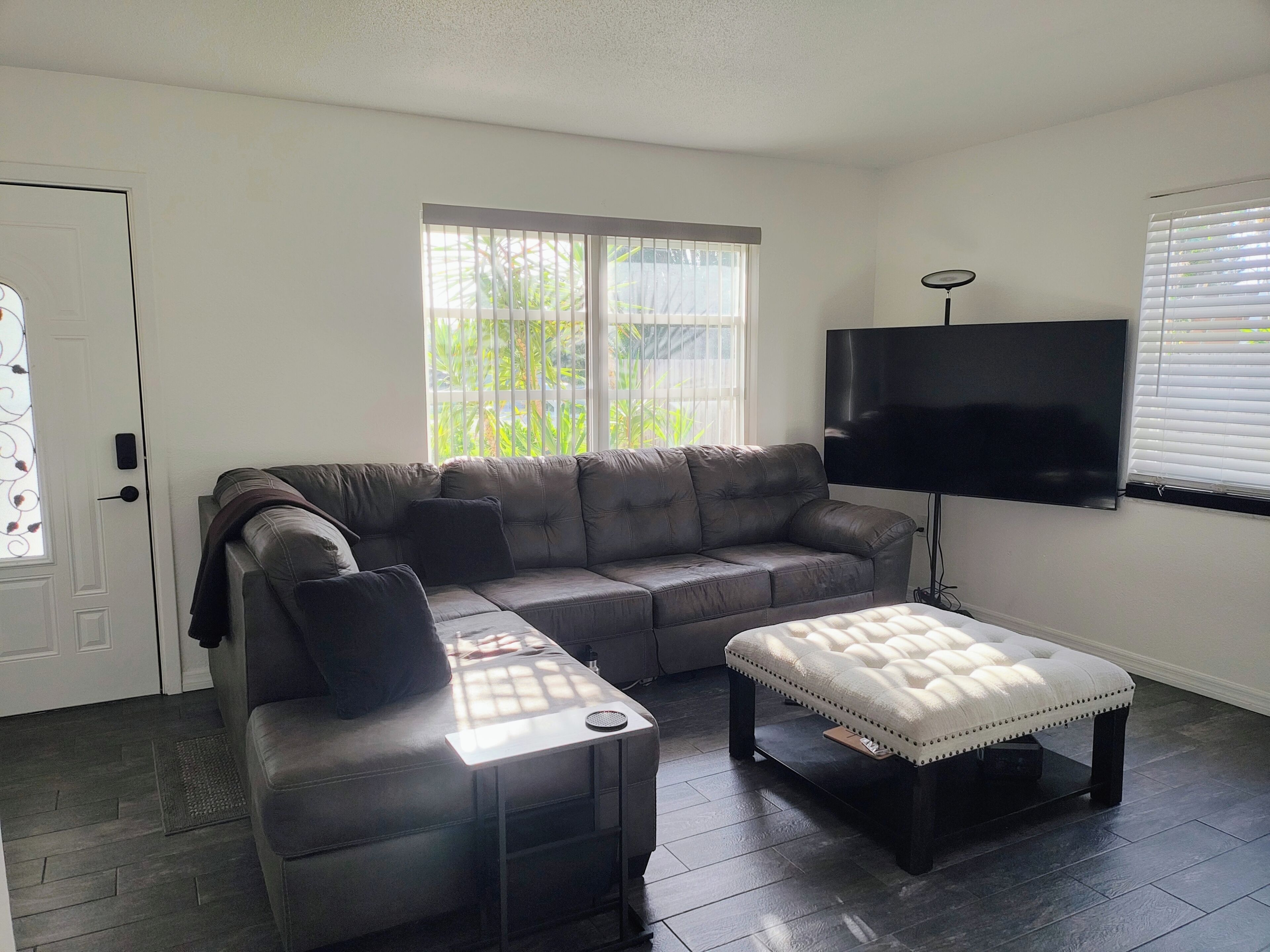 Ein Wohnzimmer mit grauem Sofa, weißem Hocker, einem Flachbildfernseher und einem Fenster mit Jalousien.