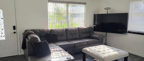 Ein Wohnzimmer mit grauem Sofa, weißem Hocker, einem Flachbildfernseher und einem Fenster mit Jalousien.