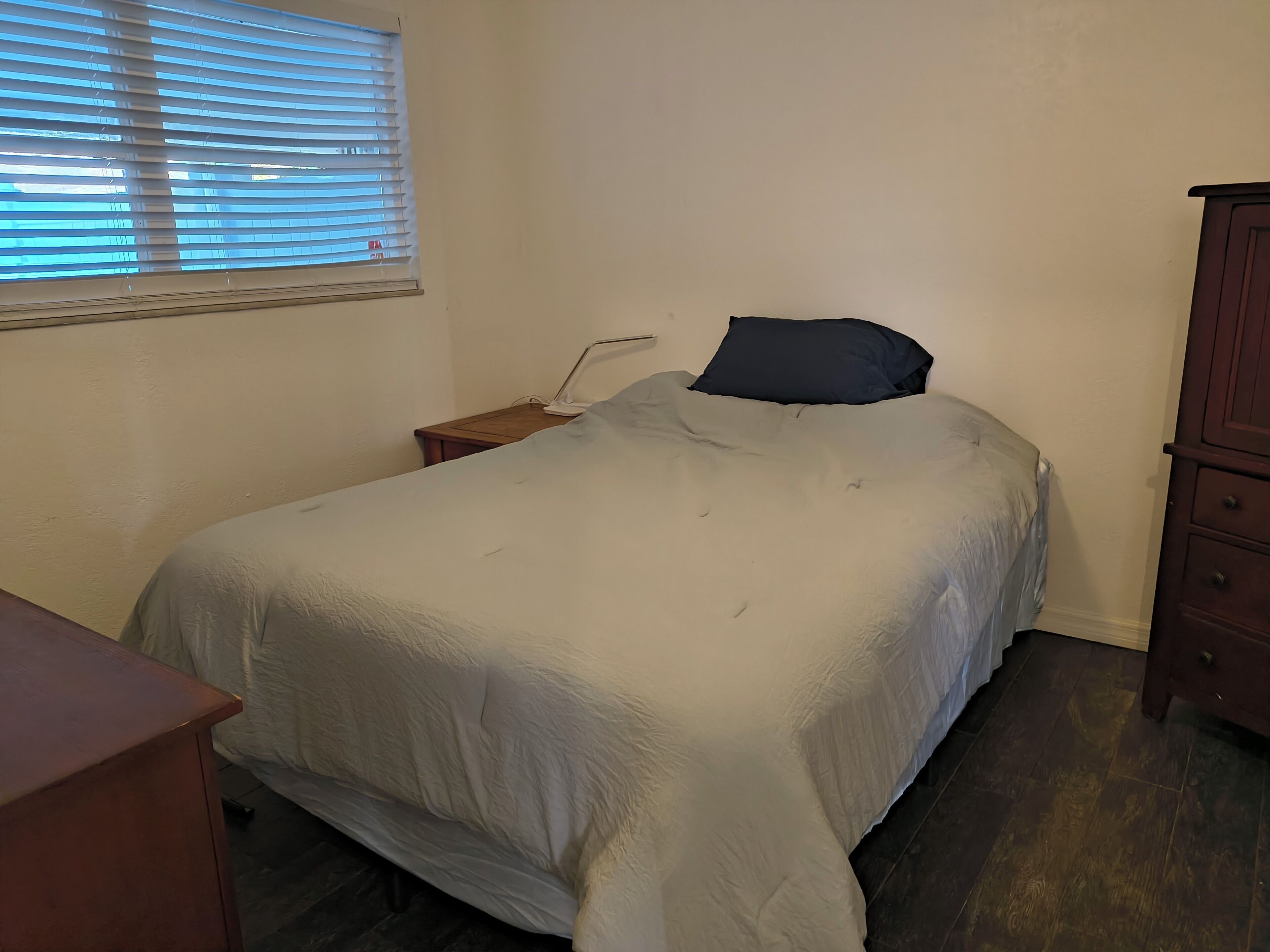 A bedroom with a bed, a nightstand, a lamp, a window with blinds, and a wooden dresser.