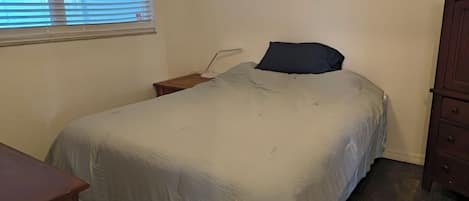 A bedroom with a bed, a nightstand, a lamp, a window with blinds, and a wooden dresser.