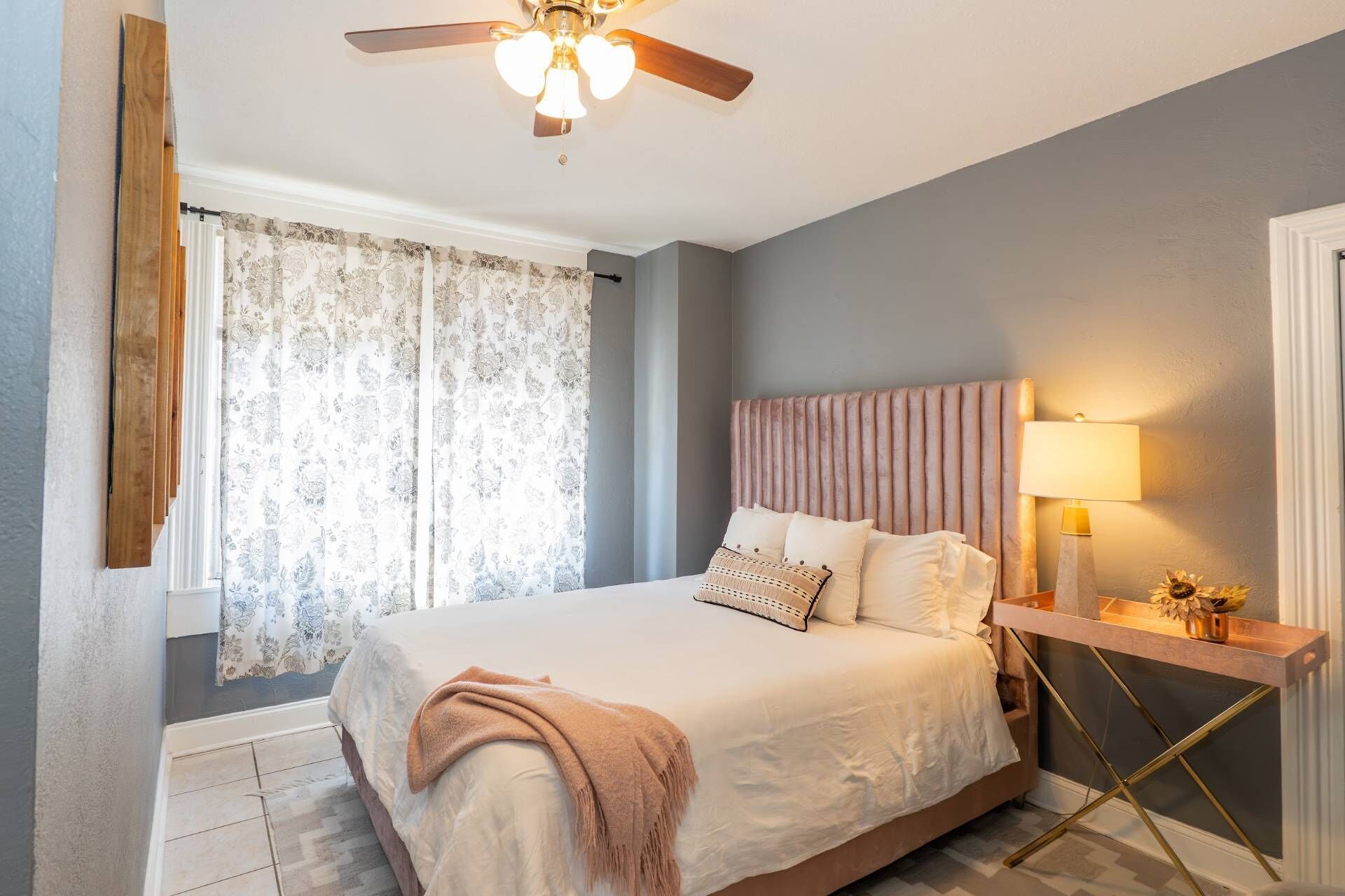 A bedroom with a bed, a ceiling fan, a bedside table with a lamp, and a window with curtains.