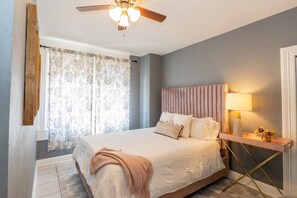 A bedroom with a bed, a ceiling fan, a bedside table with a lamp, and a window with curtains. - Casa Grande | Historic 2BR Bungalow with Private Porch Near Ybor, Armature Works & Downtown Tampa (Tampa)