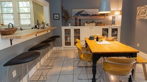 A kitchen with a wooden table, yellow chairs, a bar area with stools, and a hanging light fixture. - Casa Grande | Historic 2BR Bungalow with Private Porch Near Ybor, Armature Works & Downtown Tampa (Tampa)