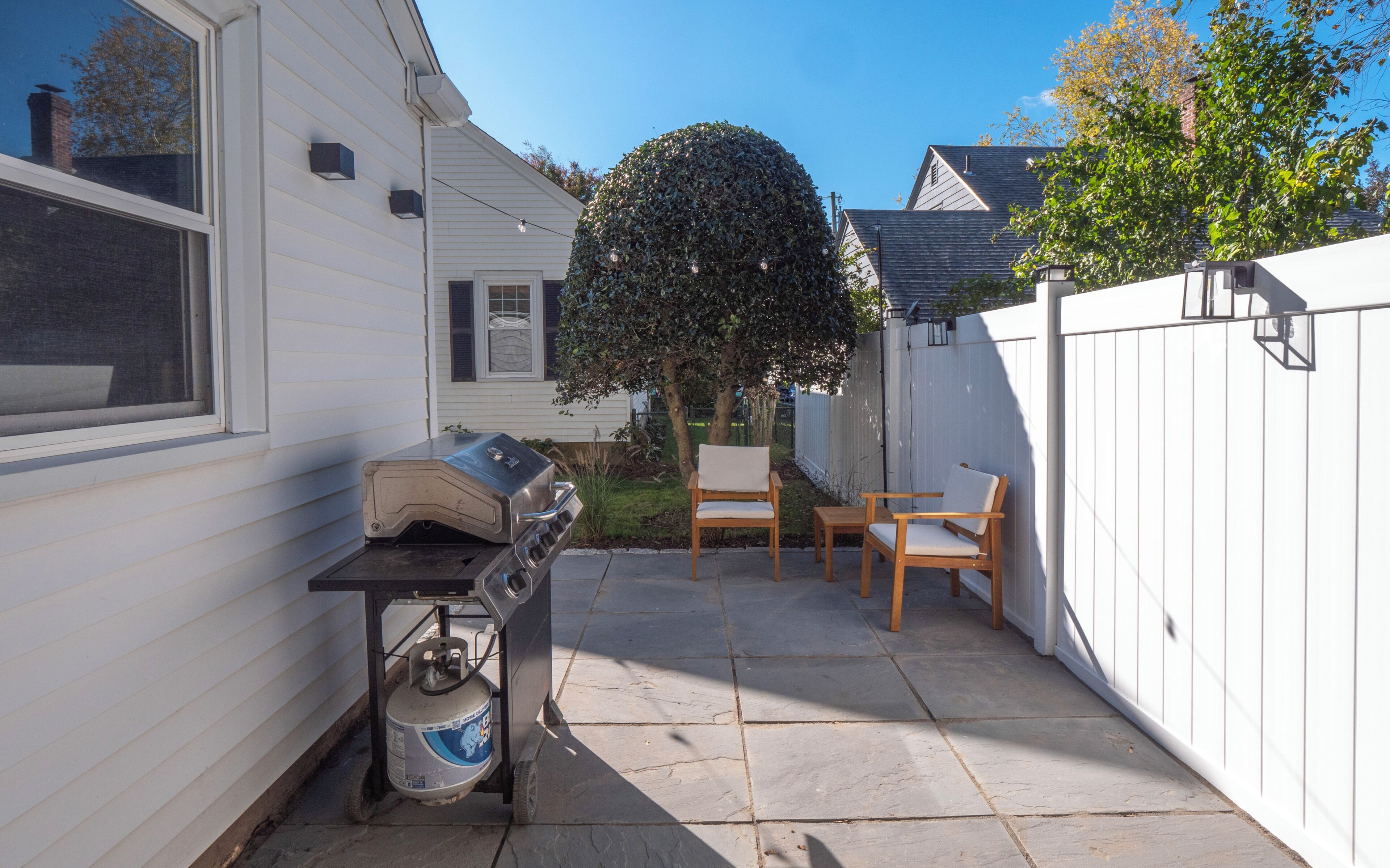 Un jardin avec un barbecue, une bouteille de gaz, deux chaises en bois et une clôture blanche.