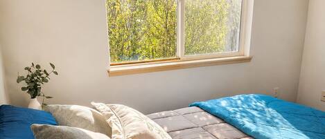 A bedroom with a bed, pillows, a window with a view of trees, and a plant on the left.