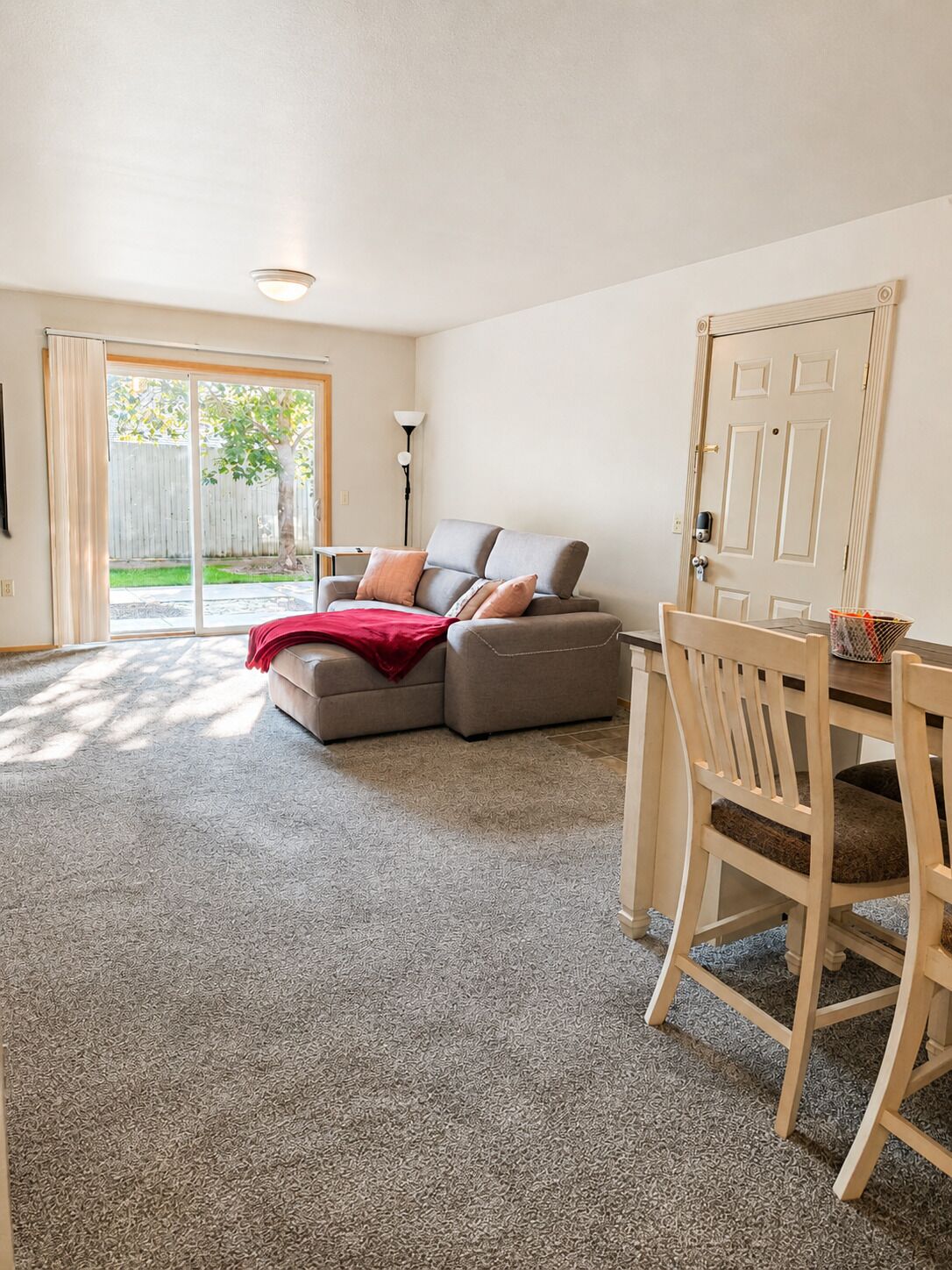 A living room with a grey sectional sofa, a red throw blanket, a wooden dining table with chairs, and a sliding glass door leading to a backyard.