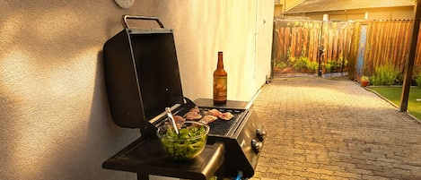 Una parrilla con carne, un bol de ensalada, una botella de cerveza y una valla de bambú al fondo.