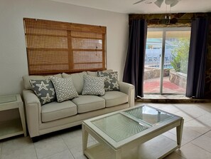 Ein Wohnzimmer mit einem beigefarbenen Sofa, gemusterten Kissen, einem Glastisch, einem gewebten Raumteiler und Blick durch ein Fenster mit Vorhängen.