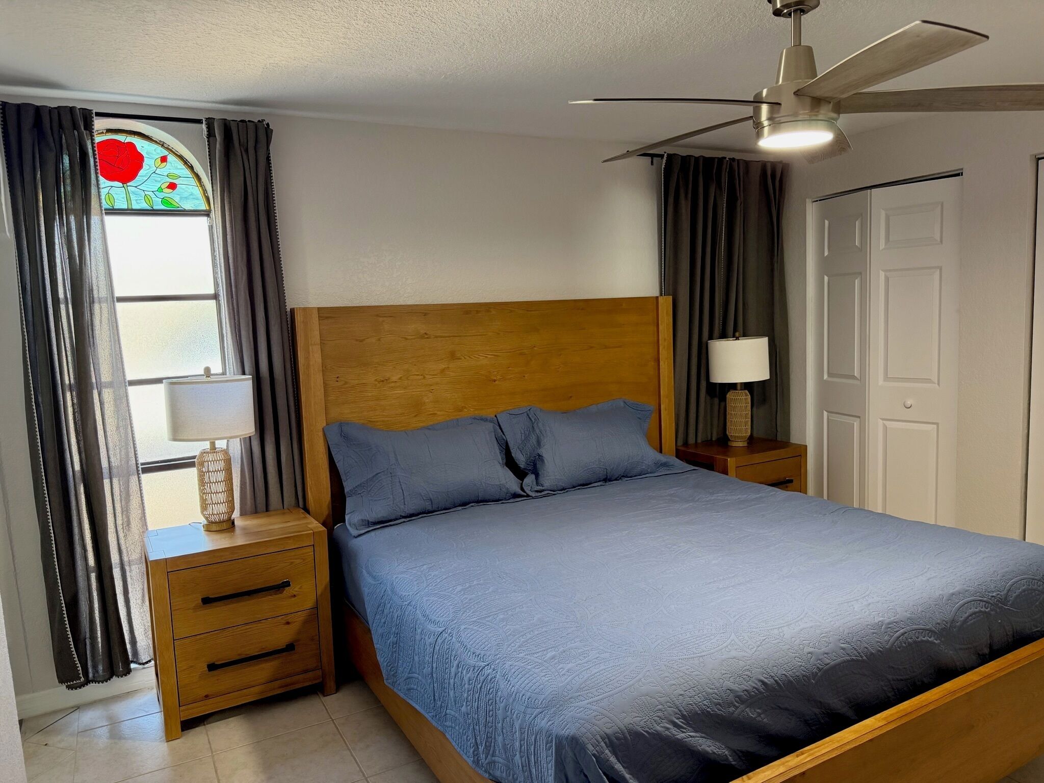 A bedroom with a wooden headboard, a single bed with a blue bedspread, a nightstand with a lamp, a window with curtains, and a ceiling fan.