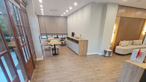 Una habitación amplia con piso de madera, un comedor con mesas y sillas, una zona de cocina y un rincón de estar con un sofá y una mesita.