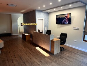 A hotel reception area with a wooden reception desk, two black office chairs, a flat-screen TV on the wall, and a staircase in the background. - Stemalu Corp  (Gjirokastër)